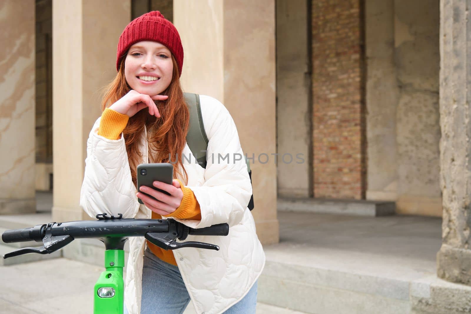 Smiling young redhead woman with mobile phone, rents e-scooter using smartphone application, riding around city, urban transport and people concept.