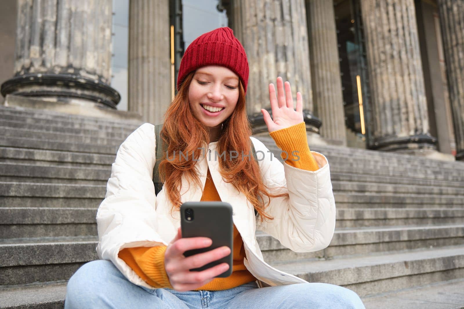 Young redhead woman sits on stairs outdoors and waves hand at smartphone camera, video chats with friends, connects to public wifi.