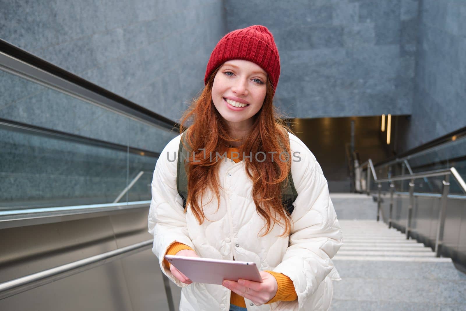 Beautiful redhead female model posing in city, walking up stairs with digital tablet, using gadget to plan her route, reading while going somewere.