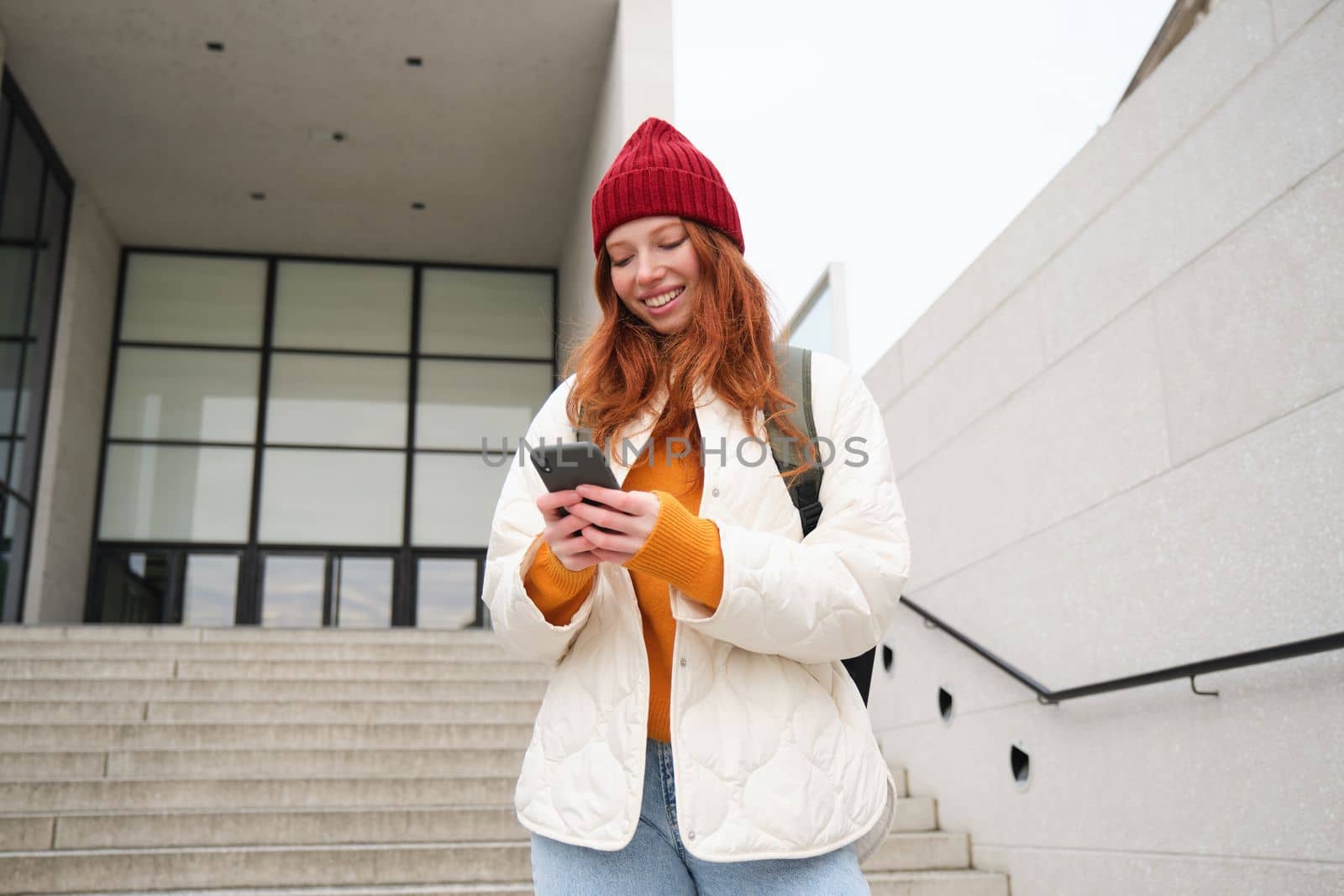 Redhead girl, young woman tourist with backpack, holds smartphone, looks for route on mobile application, searches for hotel on phone map, smiles happily.