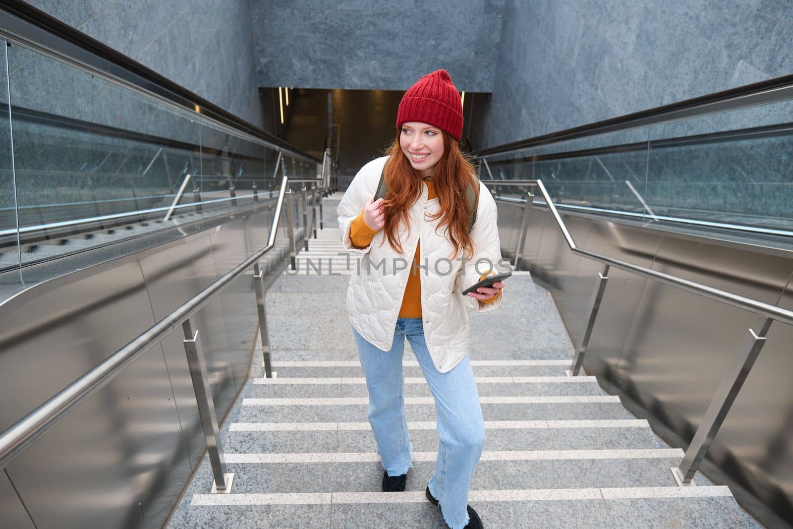 Outdoor shot of young woman plans route, follows map on smartphone app, goes up stairs with backpack and smiles.