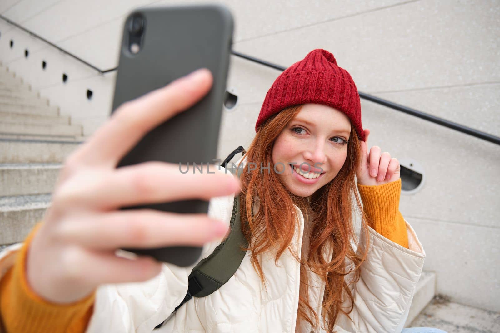 Mobile phone and people lifestyle. Stylish redhead girl takes selfie on her smartphone, poses for photo with mobile phone in hand, smiles happily, sits on stairs outdoors.