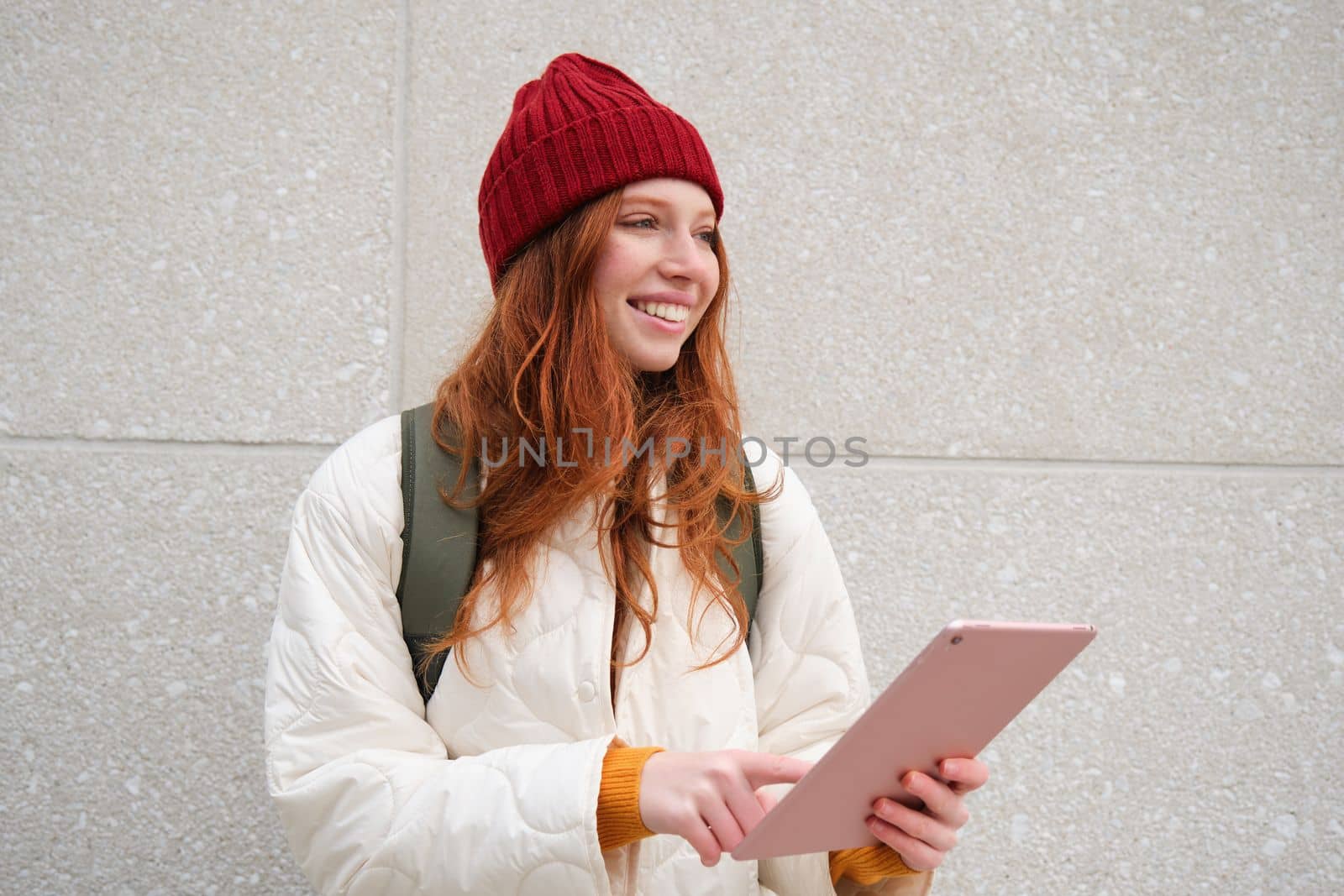 Young redhead woman with red hat, uses her digital tablet outdoors, stands on street with gadget, connects to wifi internet and searches for a location in internet.