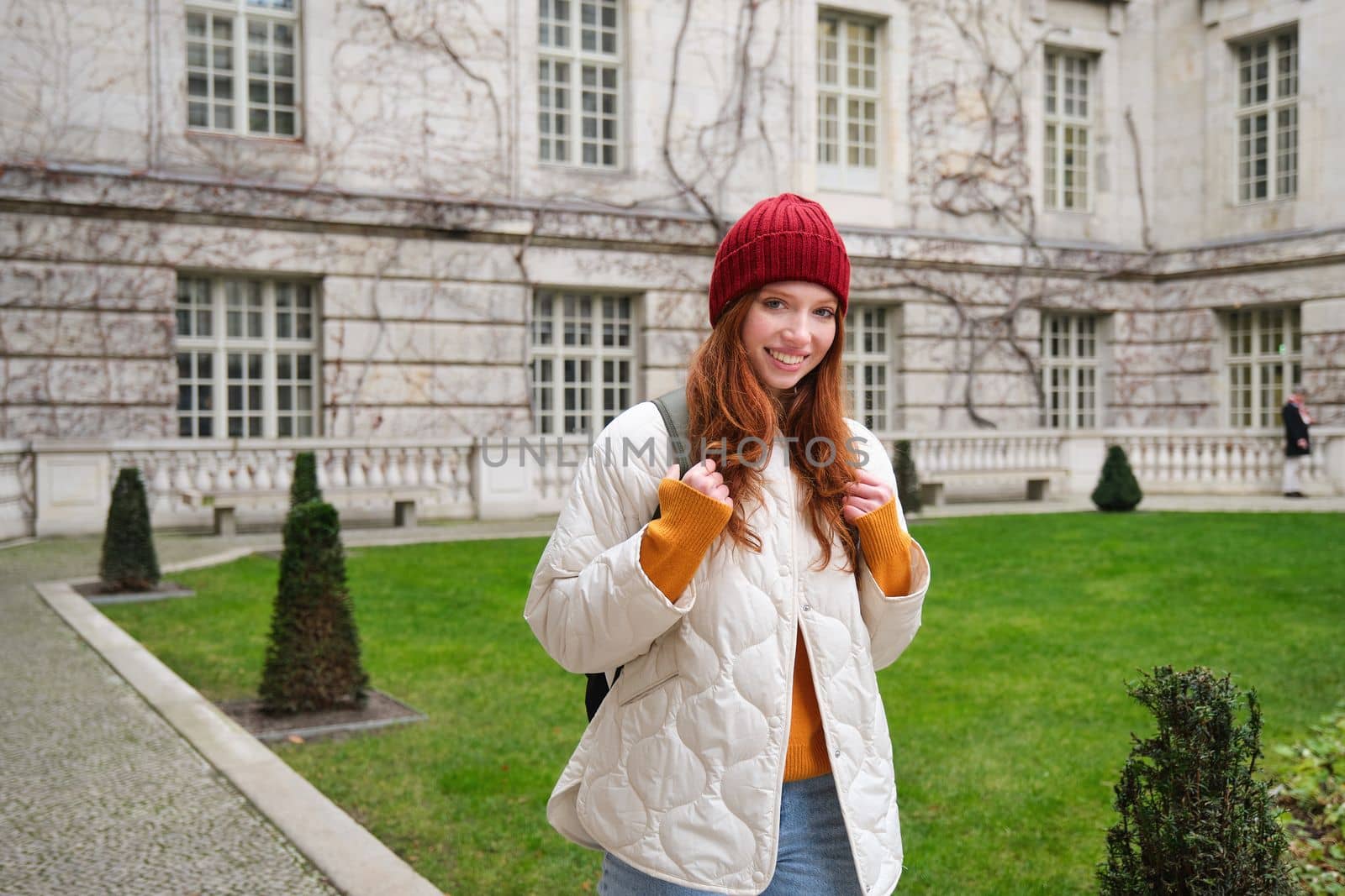 Portrait of smiling girl, backpacker in coat and red hat, wearing warm clothes for tourist trip around Europe in winter, walking around historical building.