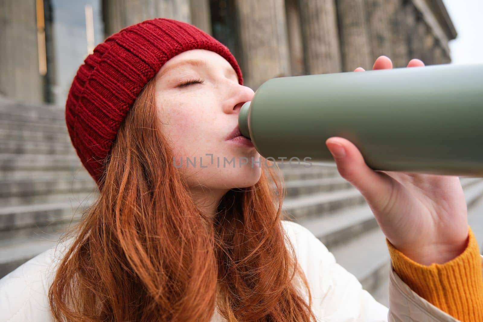 Portrait of redhead woman drinking from thermos, sitting on street stairs and enjoys hot drink from flask.