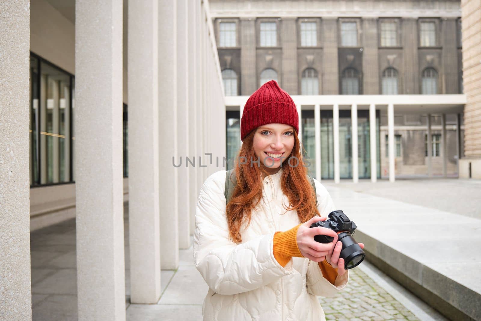 Smiling redhead girl photographer, taking pictures in city, makes photos outdoors on professional camera. Young talent and hobby concept