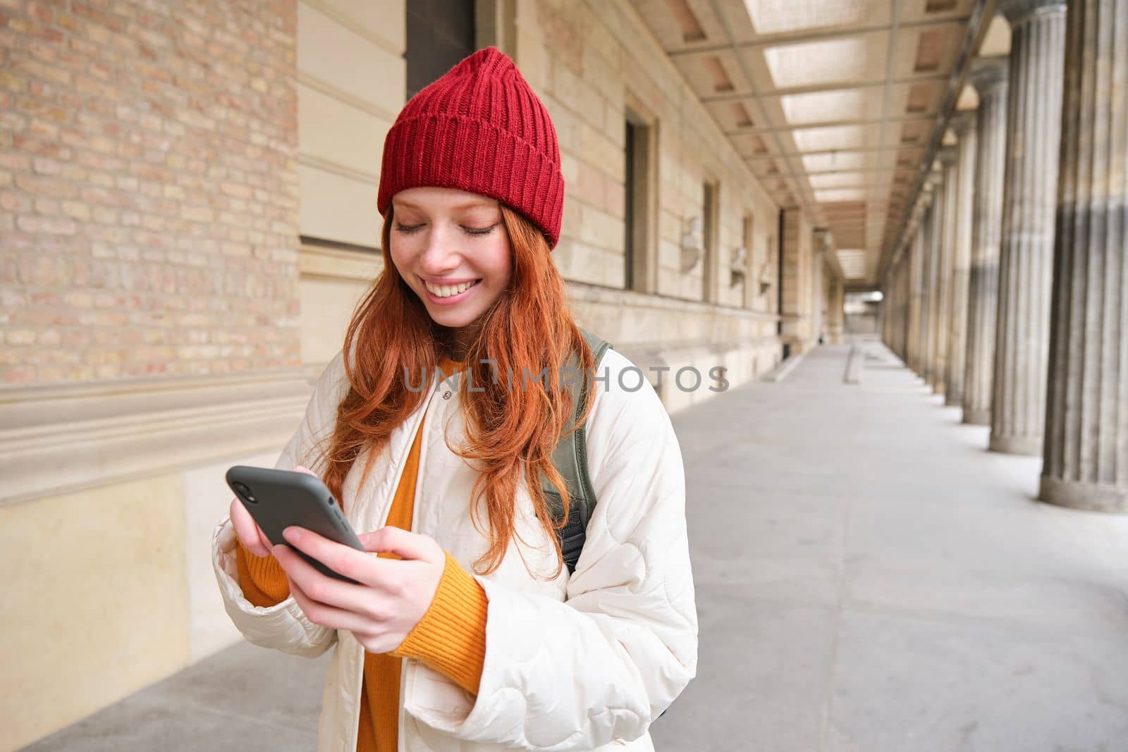 Smiling redhead girl in red hat, looks at map on smartphone app, checks her location, searches for a hostel. Tourist searches for popular attractions on mobile phone.