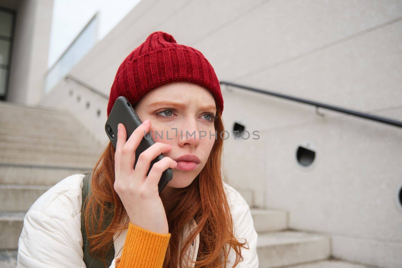 Redhead girl with concerned face, looking worried while answering phone call, hears bad news over telephone conversation, rings someone with upset emotion.