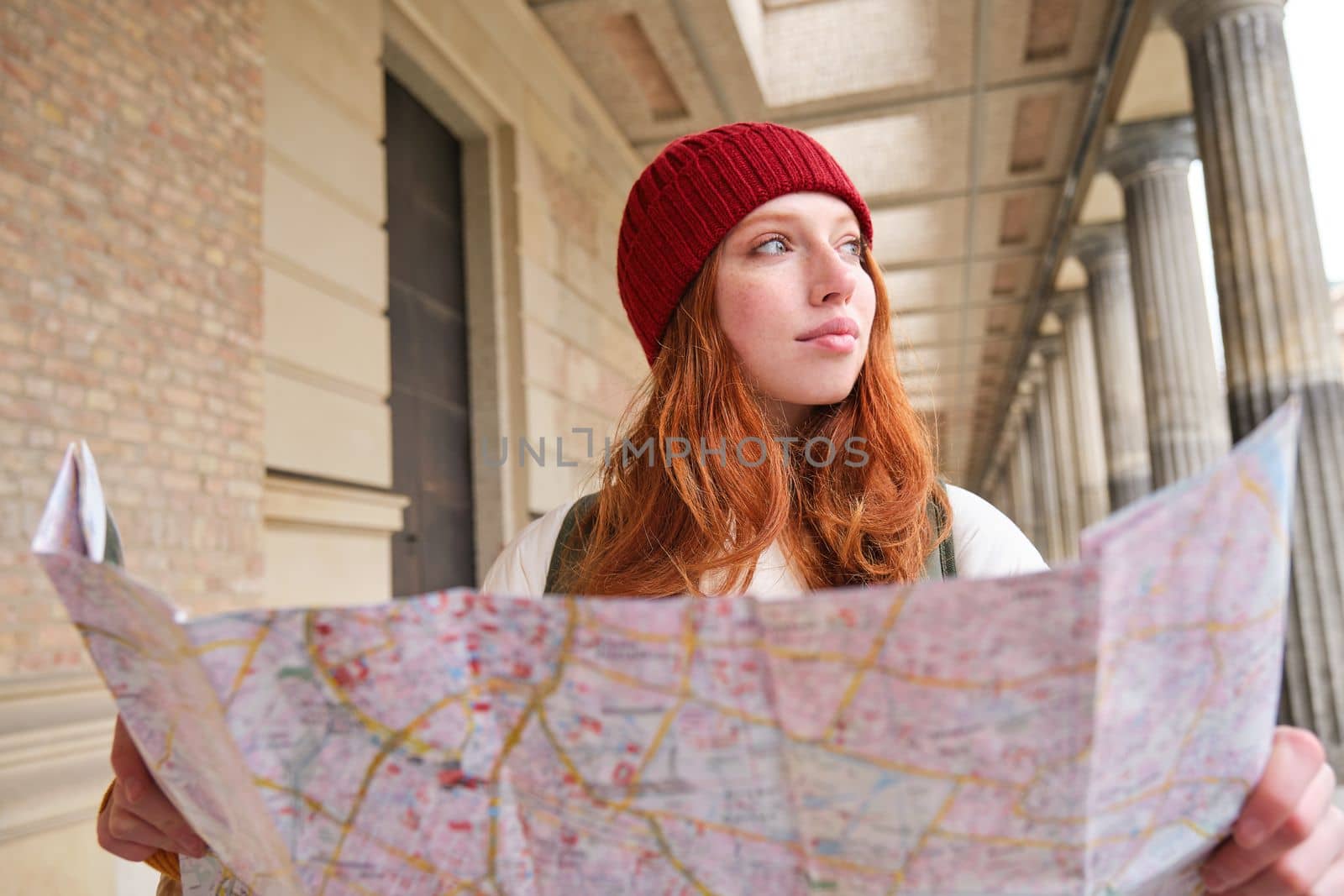 Smiling young redhead woman in red hat, looks at paper map to look for tourist attraction. Tourism and people concept. Girl explores city, tried to find way.