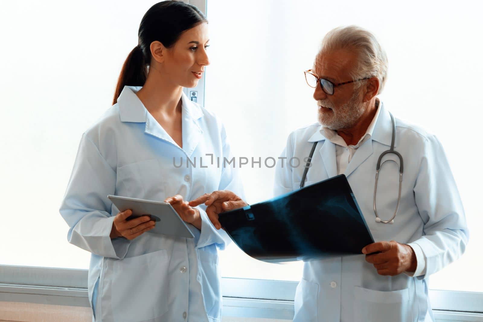 In a hospital sterile room, two professional radiographers hold and examine a radiograph for medical xray diagnosis. Novice doctor seeks advice on a patient's condition from experienced older doctor.