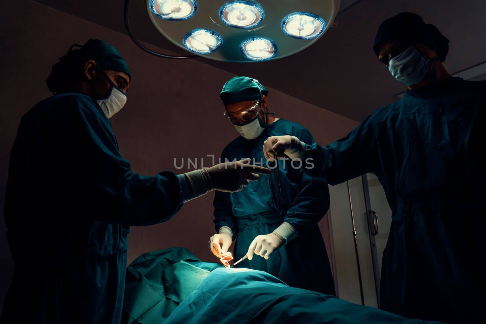 Surgical team performing surgery to patient in sterile operating room. In a surgery room lit by a lamp, a professional and confident surgical team provides medical care to an unconscious patient.