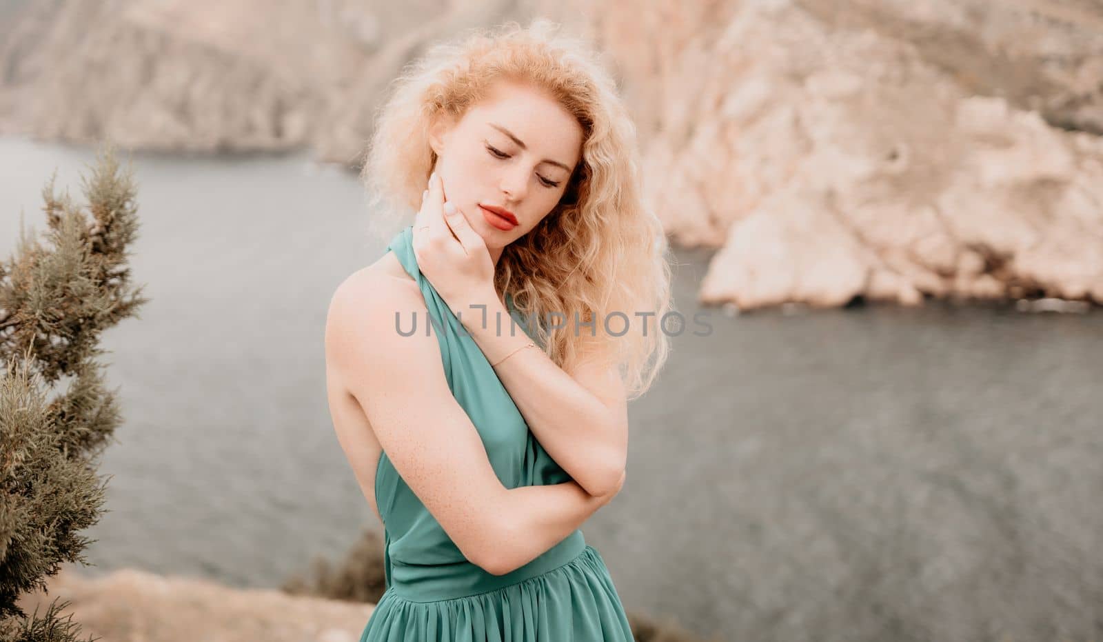 Redhead woman portrait. Curly redhead young caucasian woman with freckles looking at camera and smiling. Close up portrait cute woman in a mint long dress posing on a volcanic rock high above the sea by panophotograph