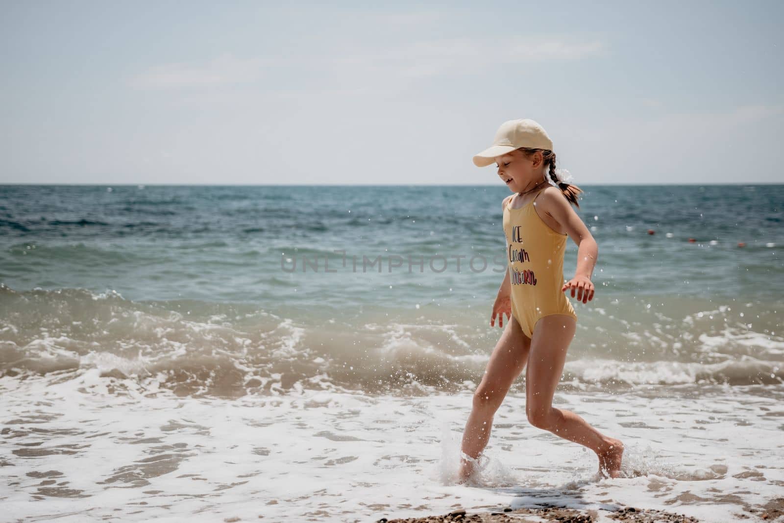Cute little girl running along the seashore against a clear blue sea and rejoices in the rays of the summer sun. Beautiful girl in yellow swimsuit running and having fun on tropical beach