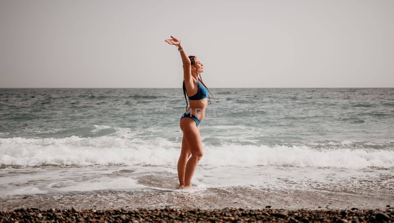 Beach vacation. Hot beautiful woman in sunhat and bikini standing with her arms raised to her head enjoying looking view of beach ocean on hot summer day.