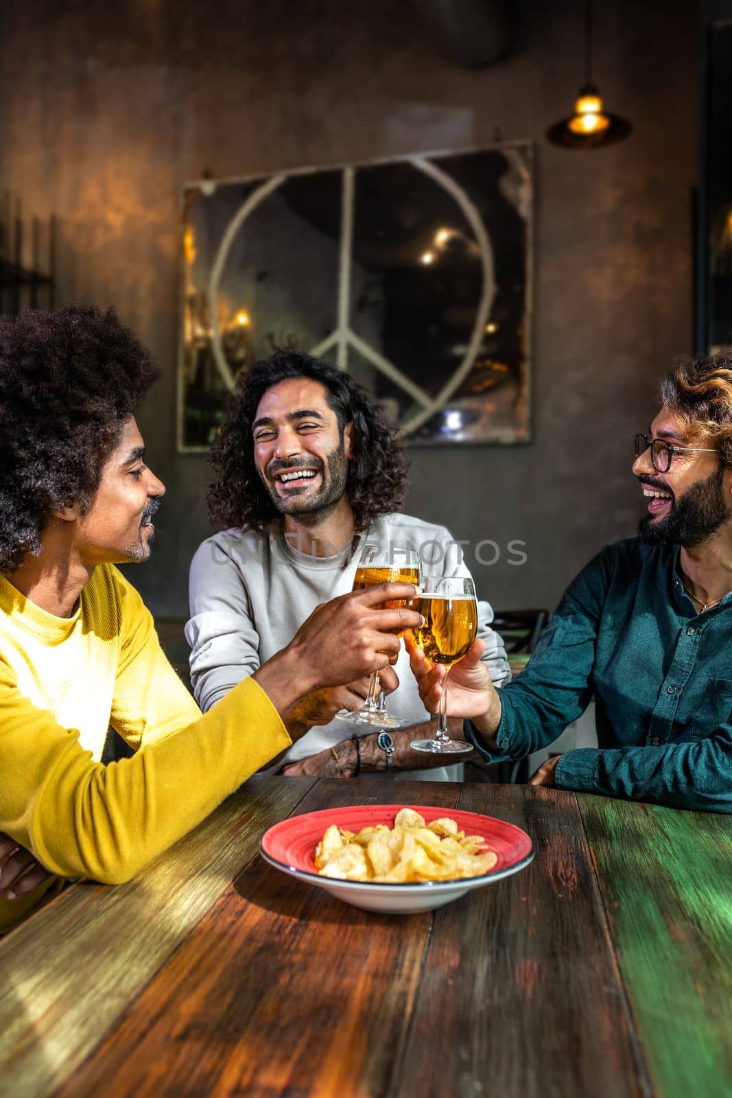 Vertical portrait of happy male friends in a bar enjoying drinks together toasting with beer, having fun. by Hoverstock