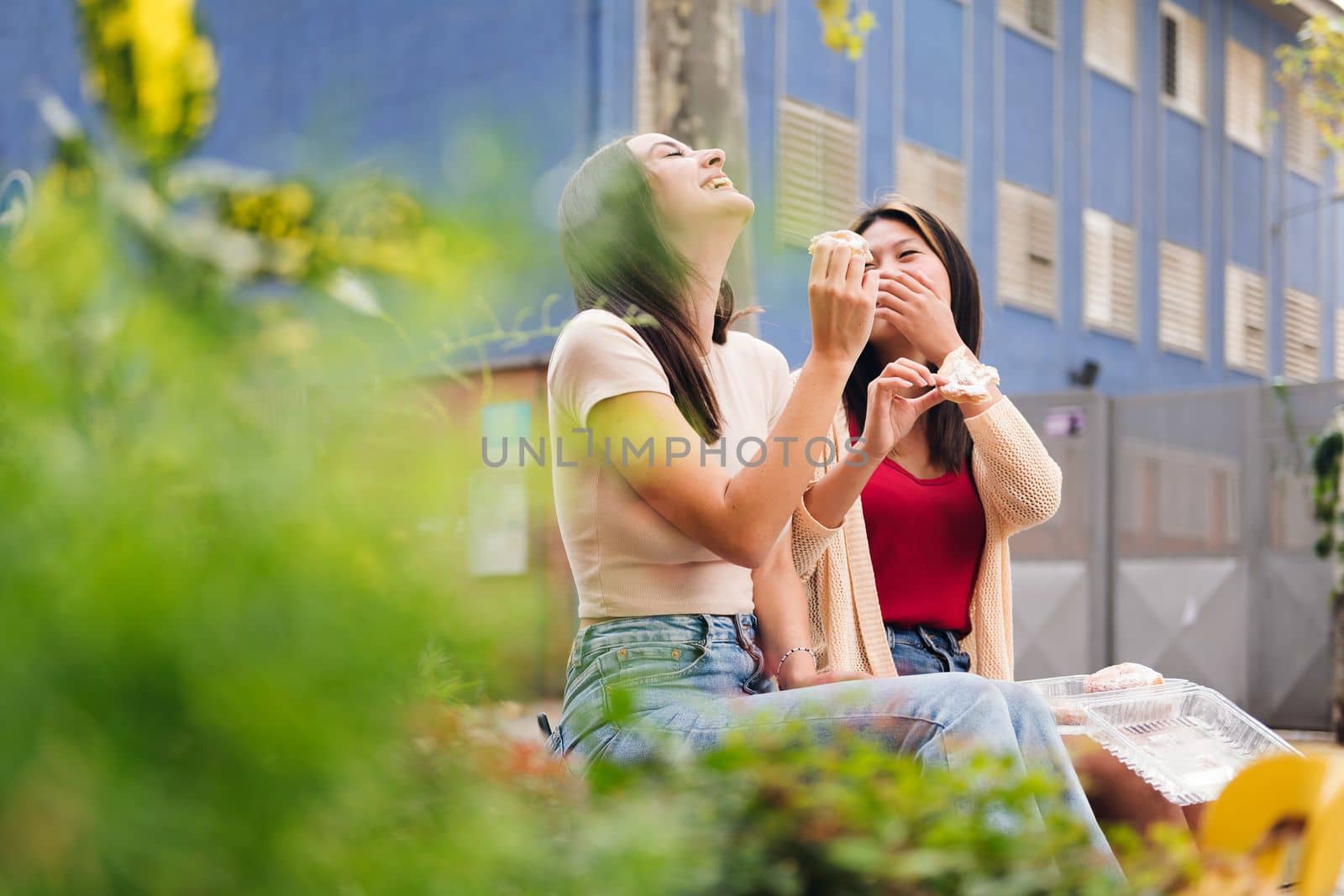 two young friends laughing and having fun sharing sweet buns while sitting in a city park, concept of friendship and love between people of the same sex