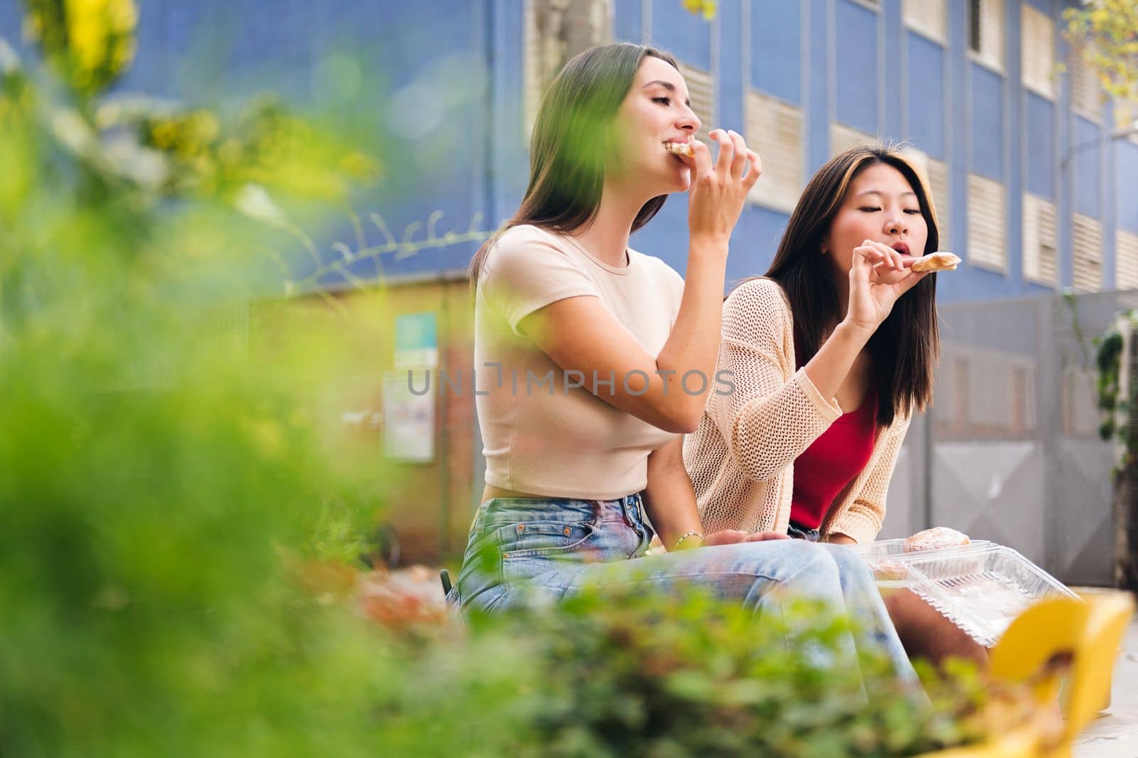 two young friends having a good time sharing some sweet buns while sitting in a city park, concept of friendship and love between people of the same sex