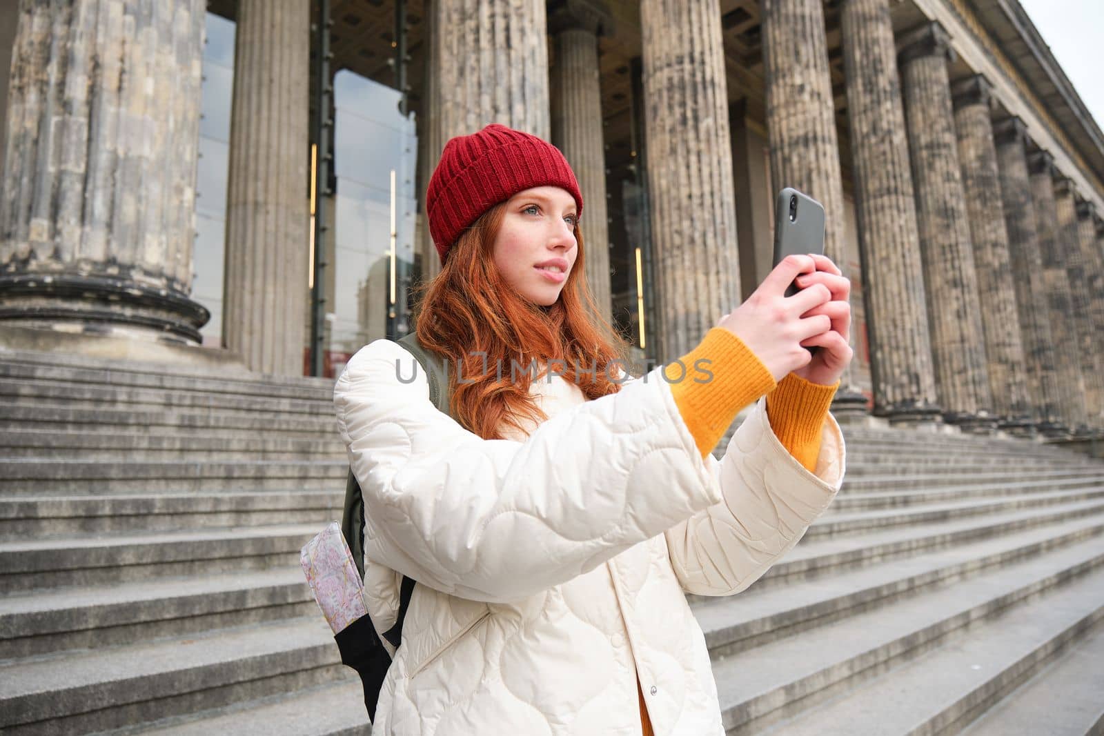 Portrait of young blogger, redhead girl tourist takes pictures of sightseeing, makes photos on smartphone camera, records video and smiles.