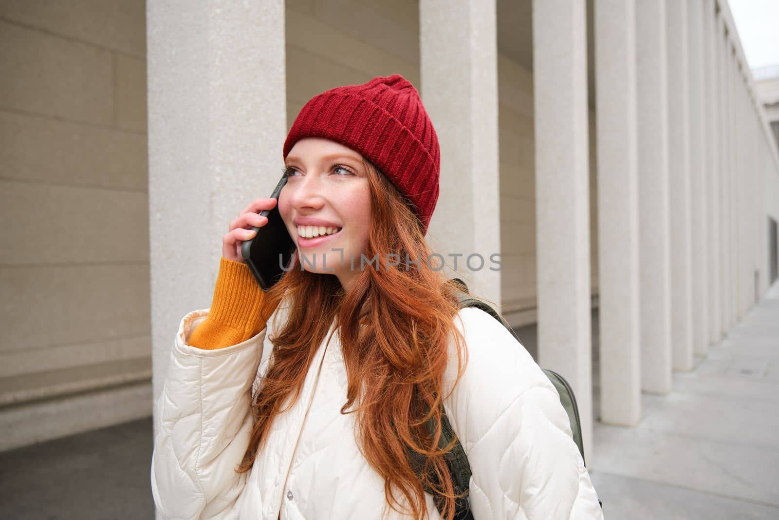 Mobile broadband and people. Smiling young redhead woman walks in town and talks on mobile phone, calling friend on smartphone, using internet to make a call abroad.