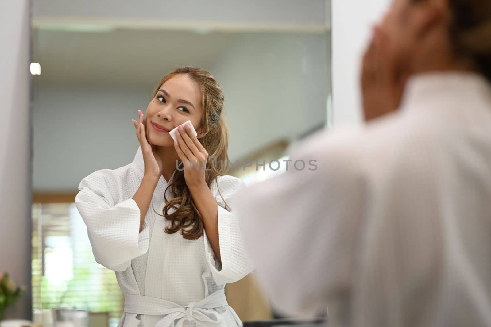 Charming young woman looking in the mirror while cleaning face with cotton pad. Beauty, skin care and lifestyle concept.