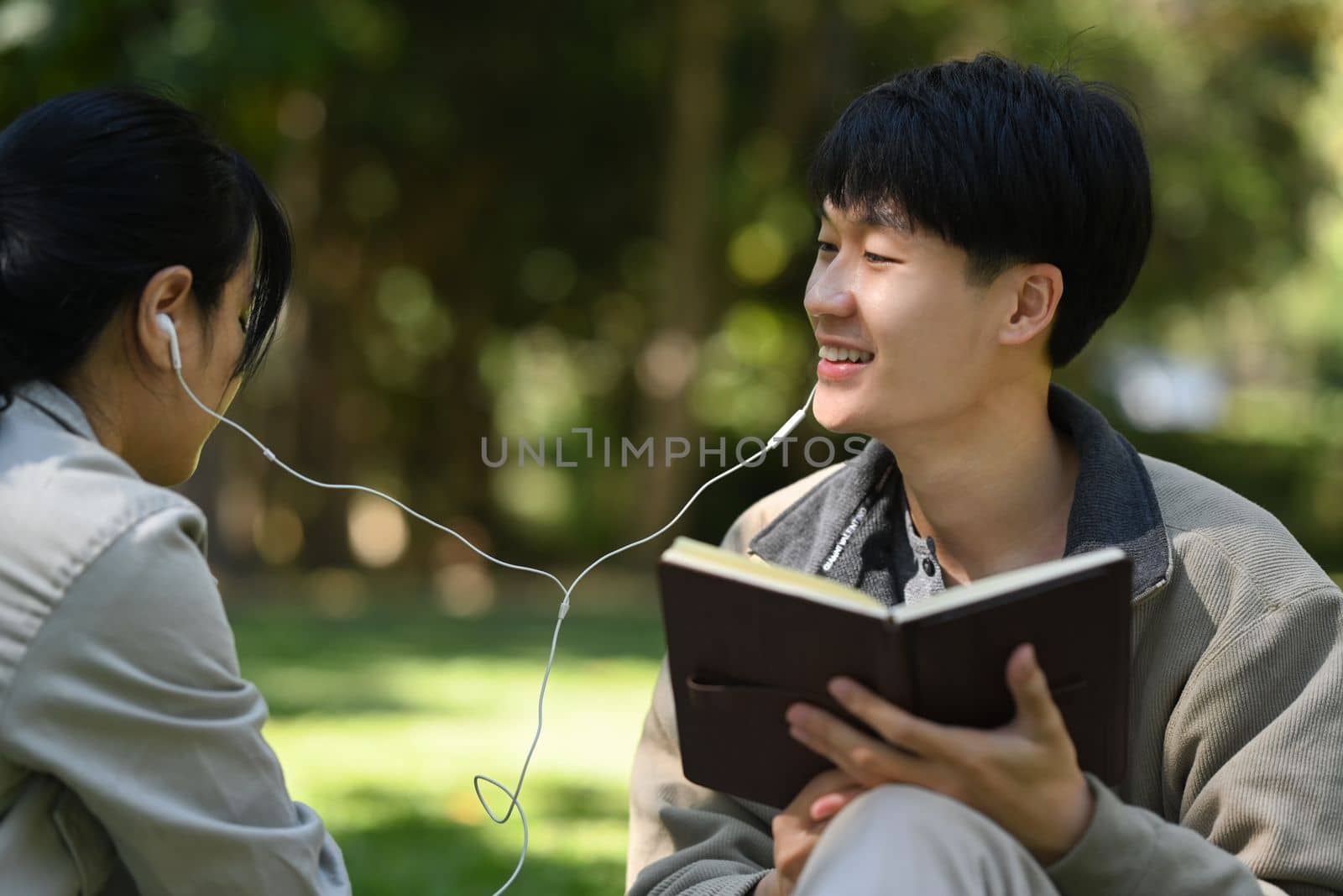 Smiling young asian man student reading a book and listening to music in earphones with his girlfriend by prathanchorruangsak