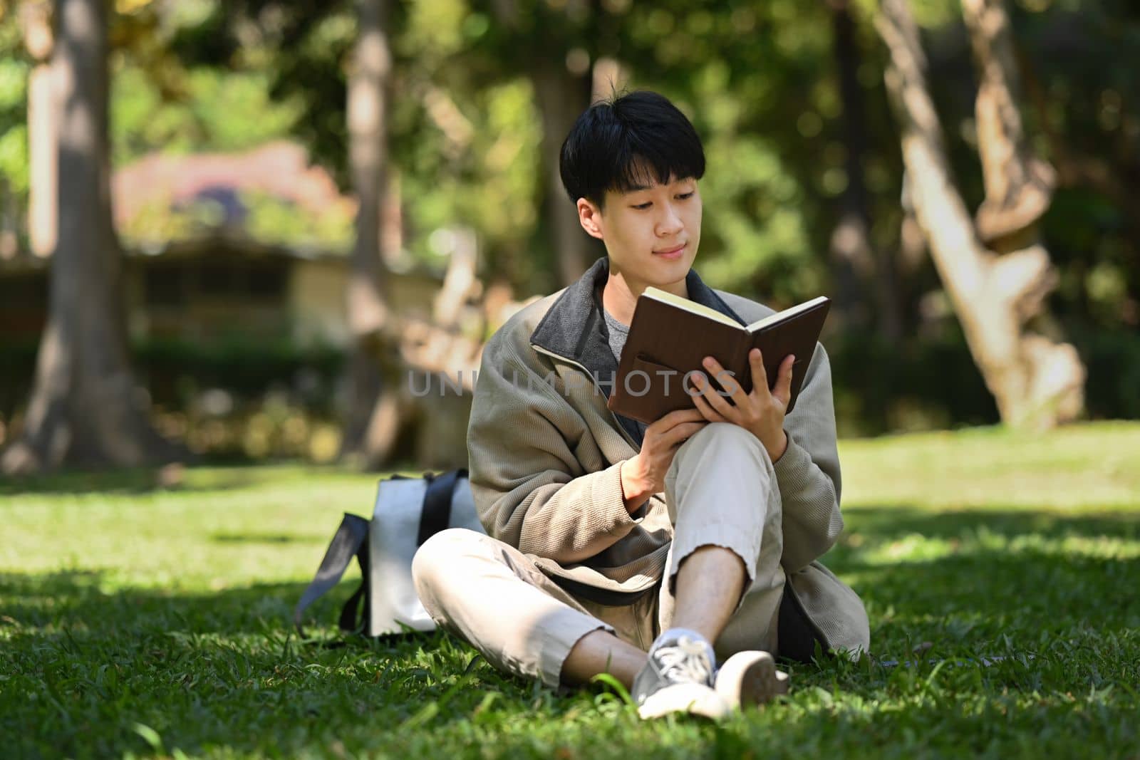 Happ young asian man reading interesting book while while sitting on green grass at sunny beautiful day.