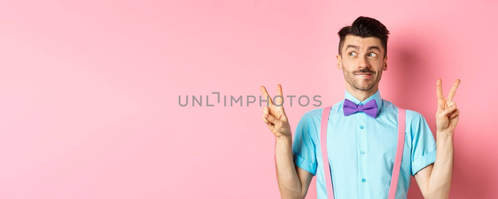 Funny young guy in bow-tie, entertain people on holiday event, showing peace signs and looking aside with excitement, standing over pink background by Benzoix