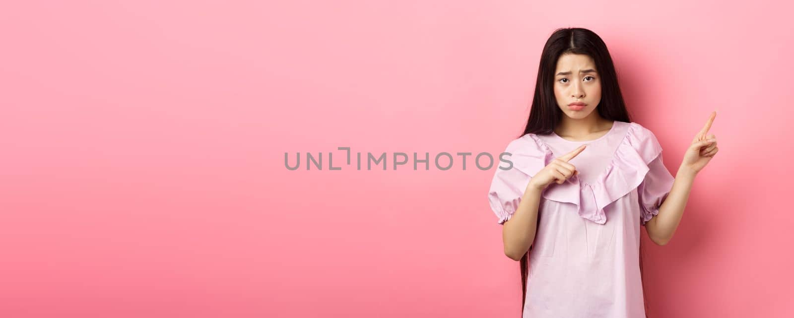 Sad and distressed asian girl frowning, sulking and pointing fingers right at disappointing bad news, feeling unhappy, standing against pink background.