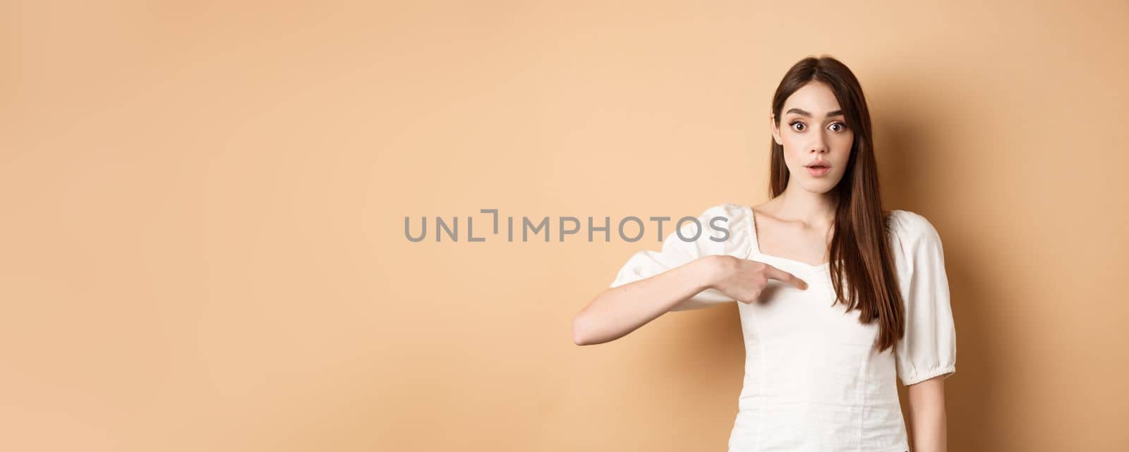 Confused girl pointing at herself with disbelief, being chosen or picked, standing on beige background by Benzoix