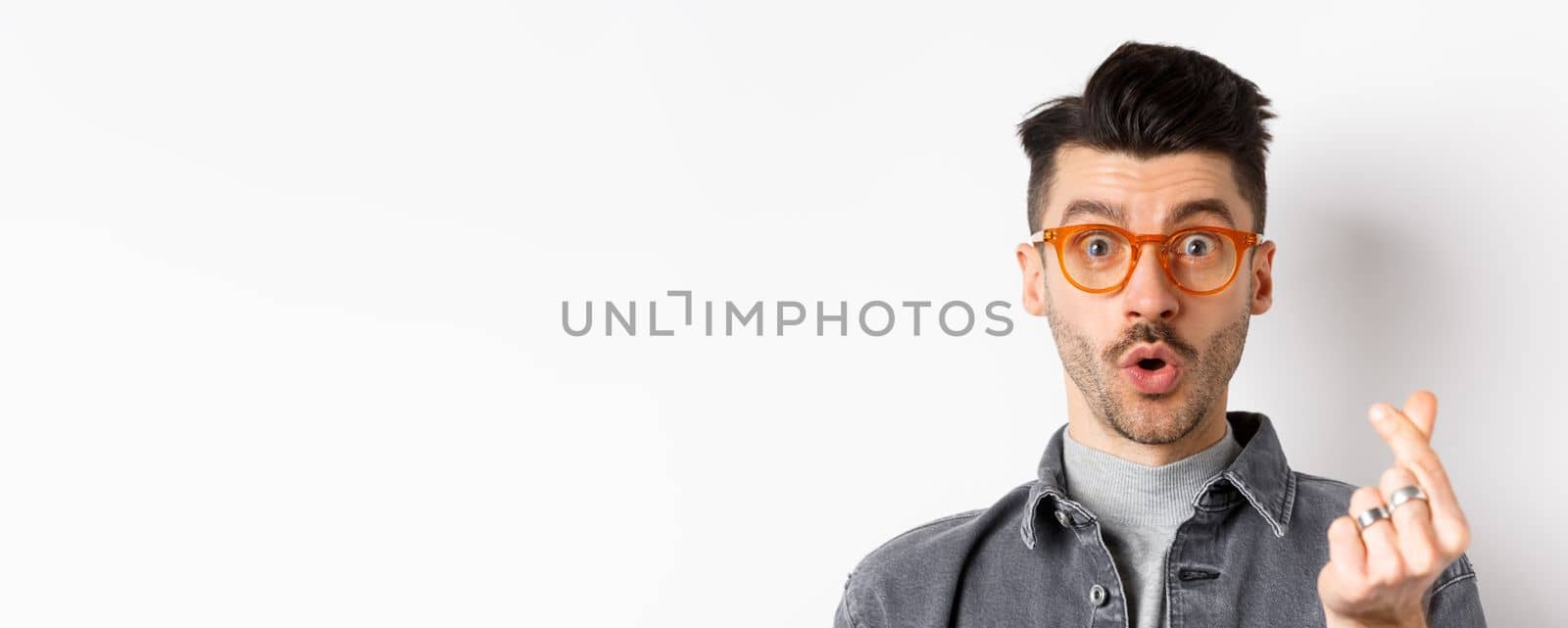 Close up portrait of cute guy with moustache wearing glasses, showing hand heart sign, look impressed at camera, standing on white background.
