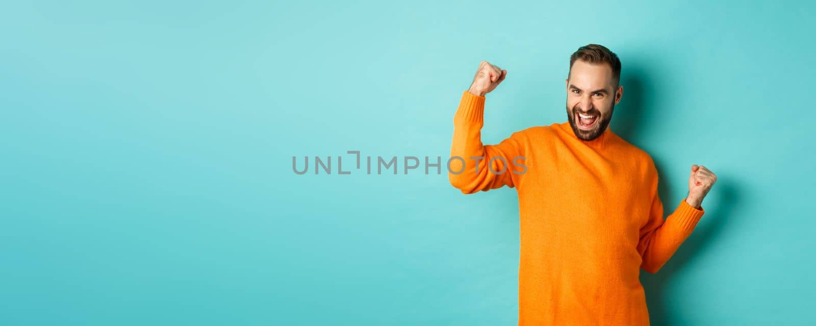 Winner. Excited man triumphing, making fist pump and saying yes, triumphing and looking satisfied, achieve goal, standing over light blue background by Benzoix