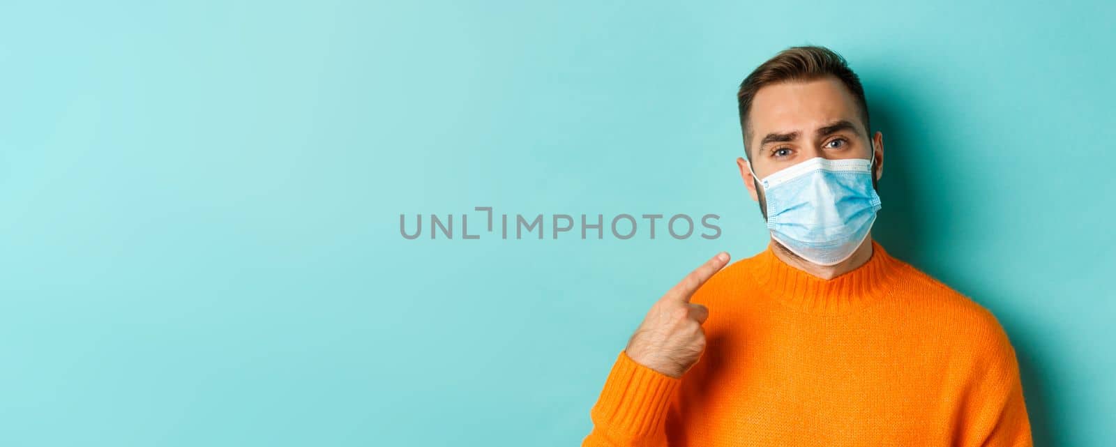 Covid-19, social distancing and quarantine concept. Upset man in complaining on face mask, pointing at it and looking bothered, standing over light blue background by Benzoix
