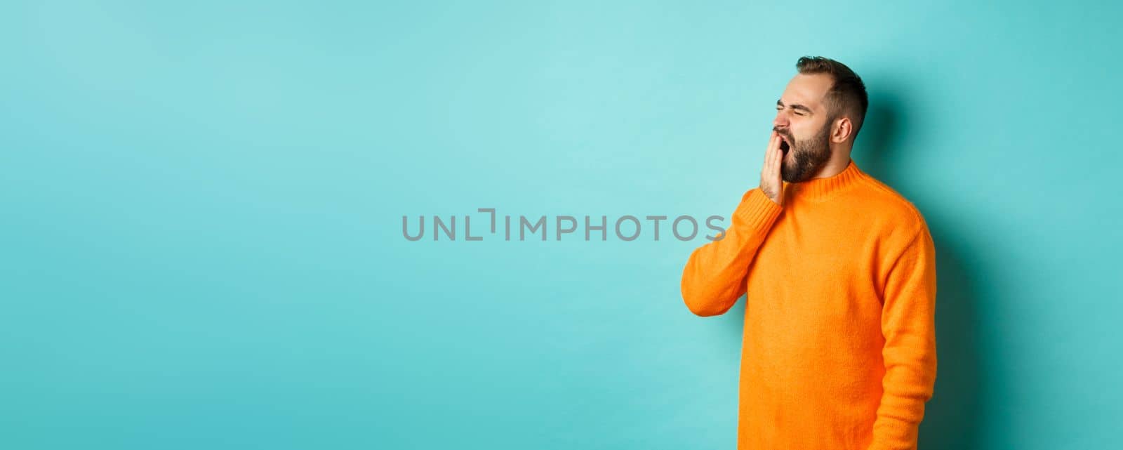 Photo of tired bearded man yawning, cover mouth with hand, standing sleepy against light blue background.
