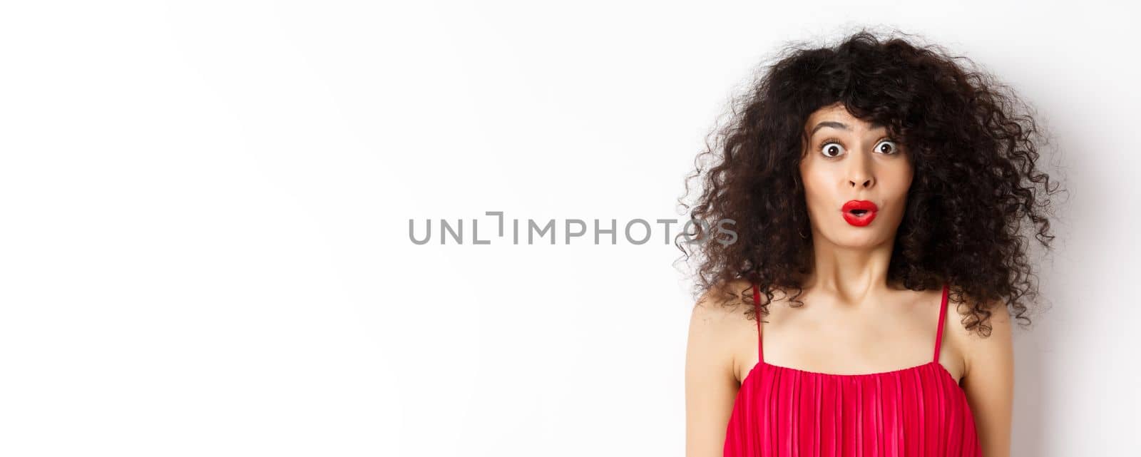 Close-up of surprised caucasian woman with red lips and curly hairstyle, saying wow and looking amazed, standing in red dress over white background by Benzoix