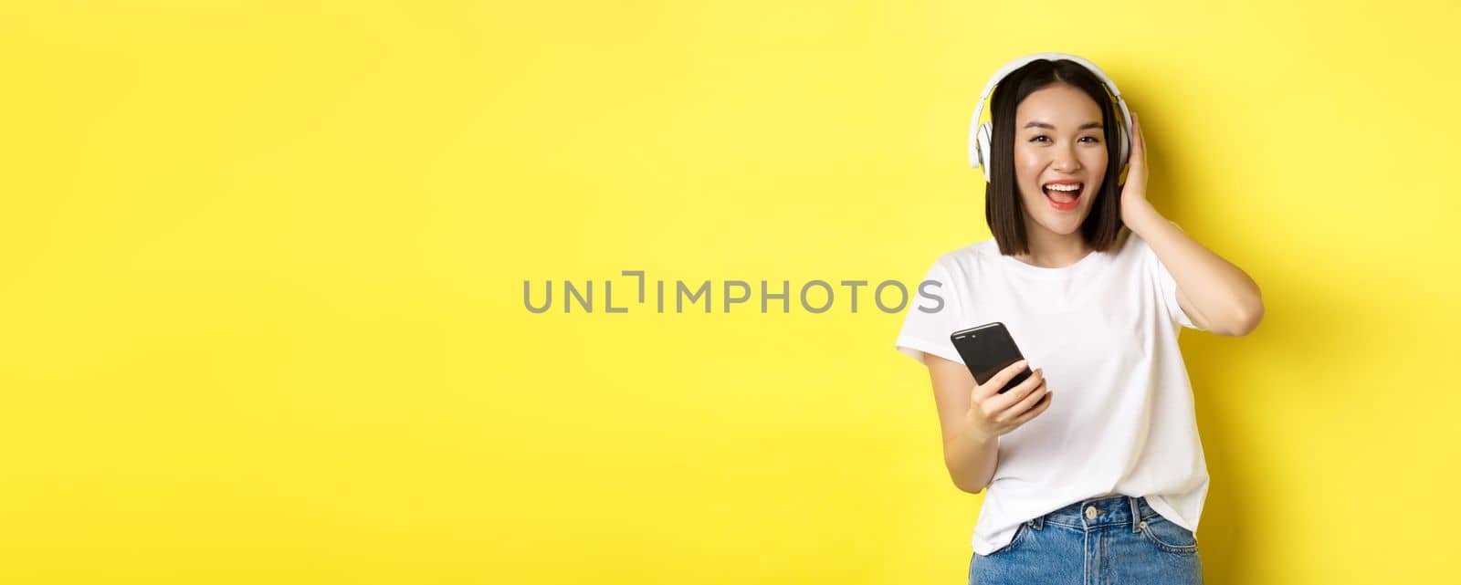 Cool asian girl dancing and listening music in wireless headphones, holding smartphone in hand, standing over yellow background.