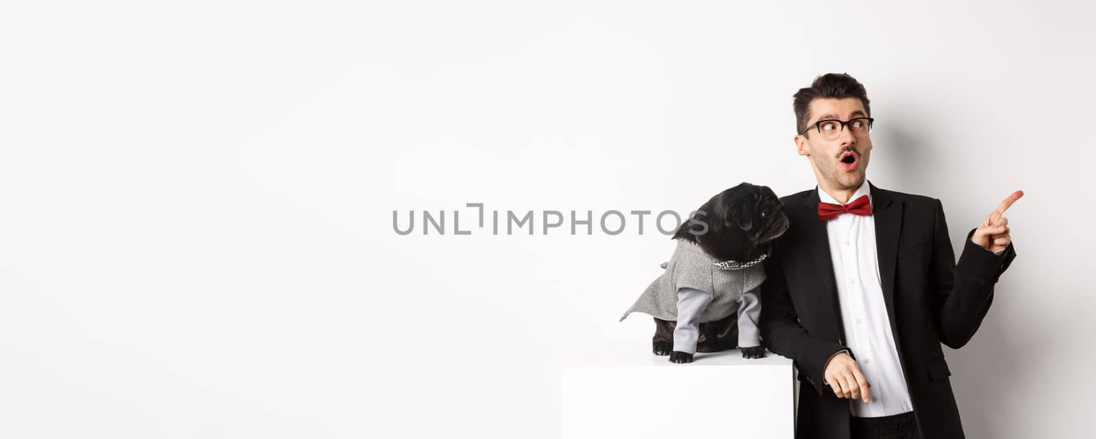 Animals, party and celebration concept. Amazed young man and black dog in costumes staring right at copy space, standing against white background.