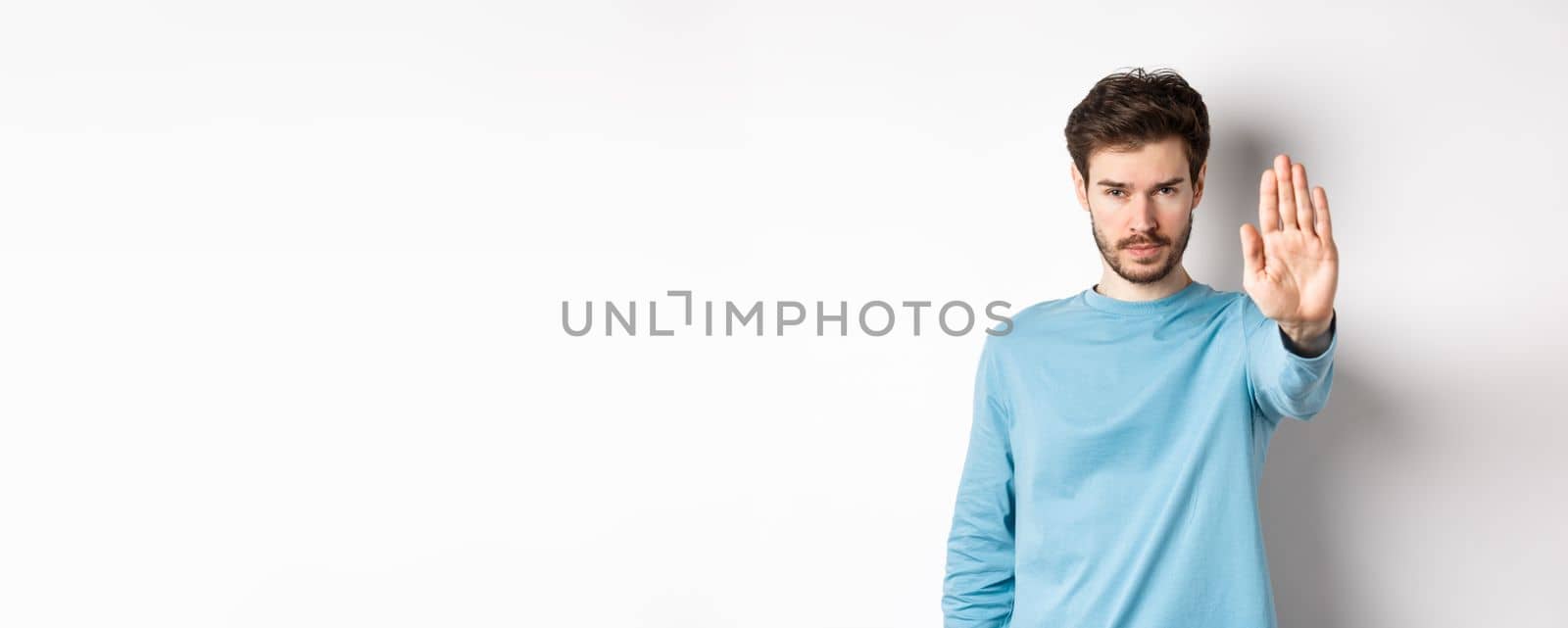 Serious young man looking confident with hand stretched out, saying stop, prohibit something bad or give warning, standing over white background by Benzoix