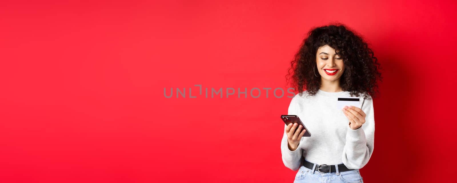 E-commerce and online shopping concept. Young modern woman paying with credit card, making purchase with smartphone, standing on red background.