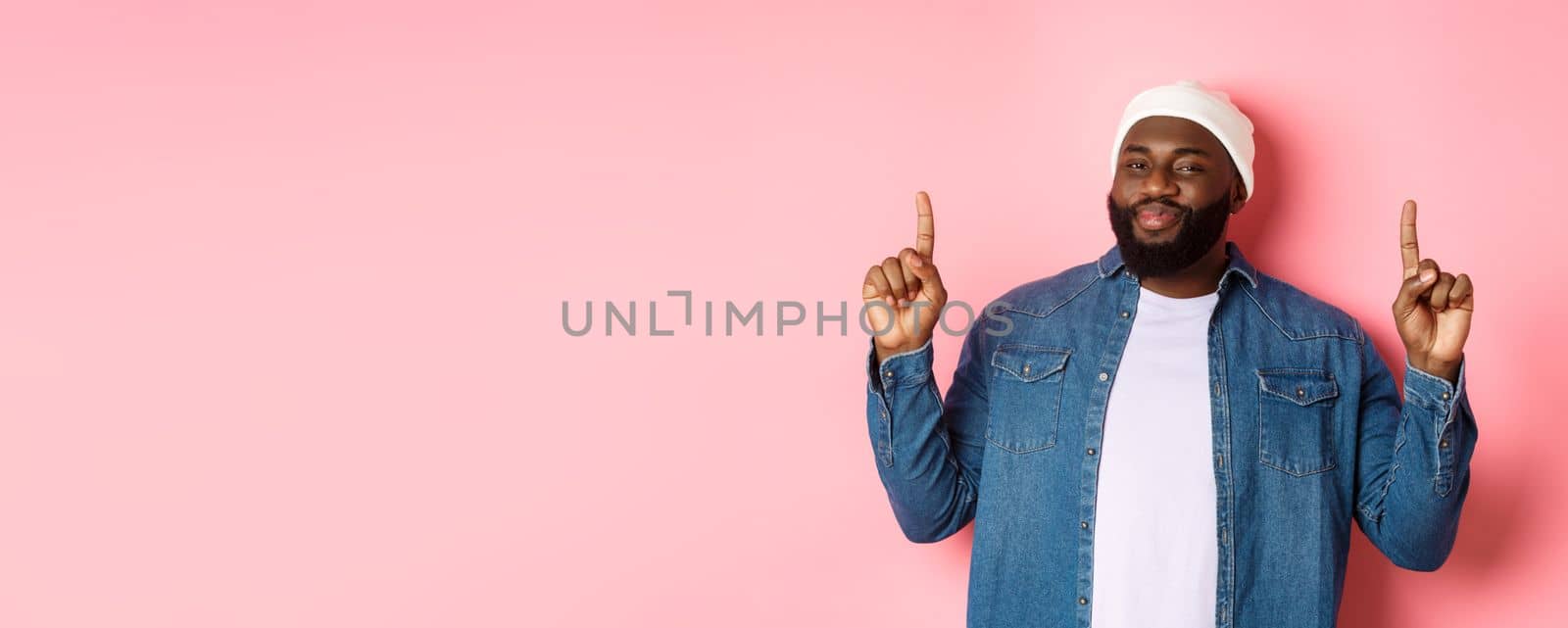 Pleased handsome Black man nodding in approval and checking out good promo, pointing fingers up at offer, showing best deal, pink background by Benzoix