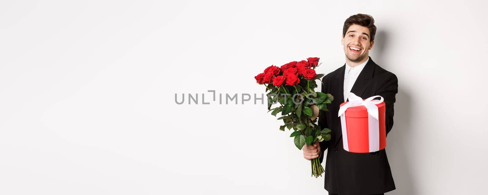 Concept of holidays, relationship and celebration. Image of handsome smiling guy in black suit, holding bouquet of red roses and giving you a gift, standing against white background by Benzoix