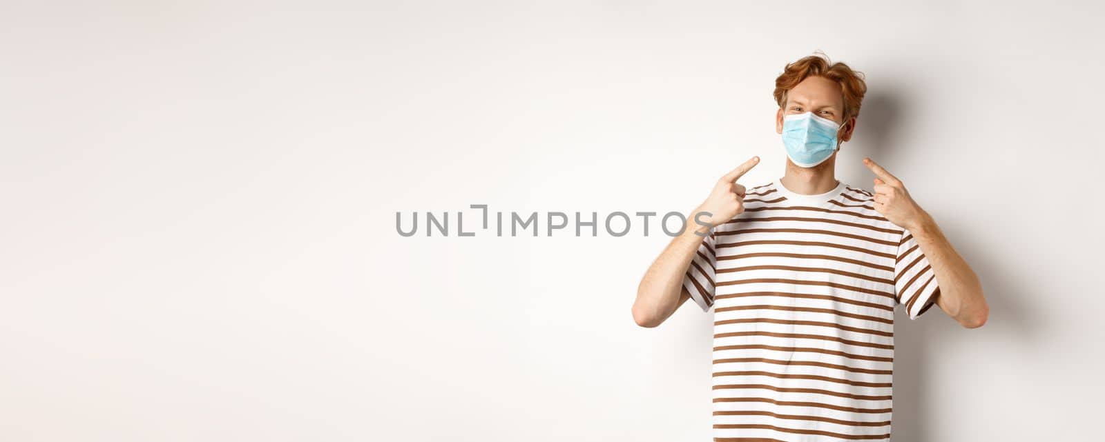 Covid, virus and social distancing concept. Smiling redhead guy pointing fingers at face mask, standing over white background by Benzoix