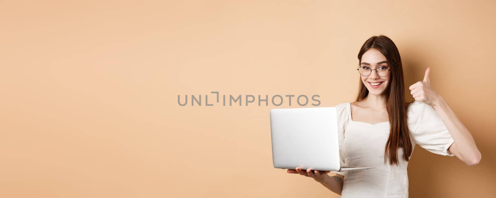 Smiling woman in glasses using laptop and showing thumb up, working on computer, standing on beige background.