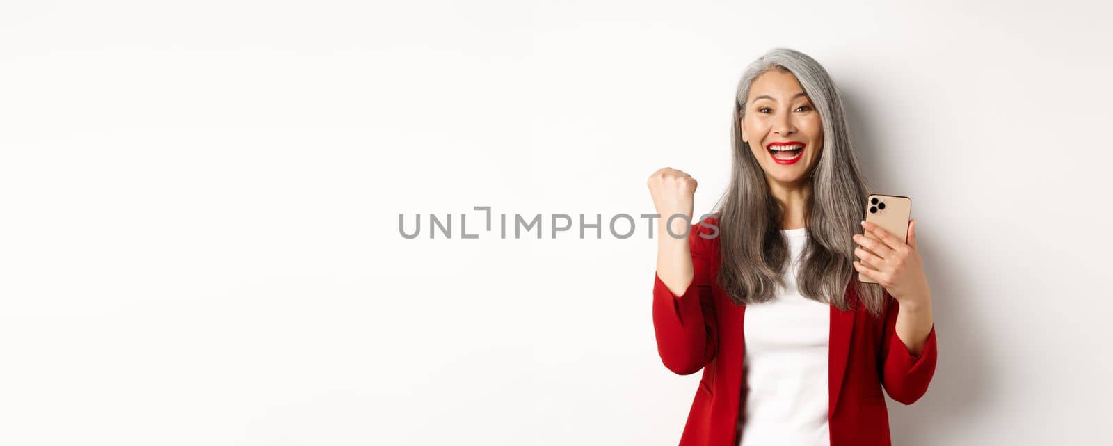 Asian old woman winning online, holding smartphone and making fist pump gesture to celebrate win, triumphing and smiling, standing over white background.