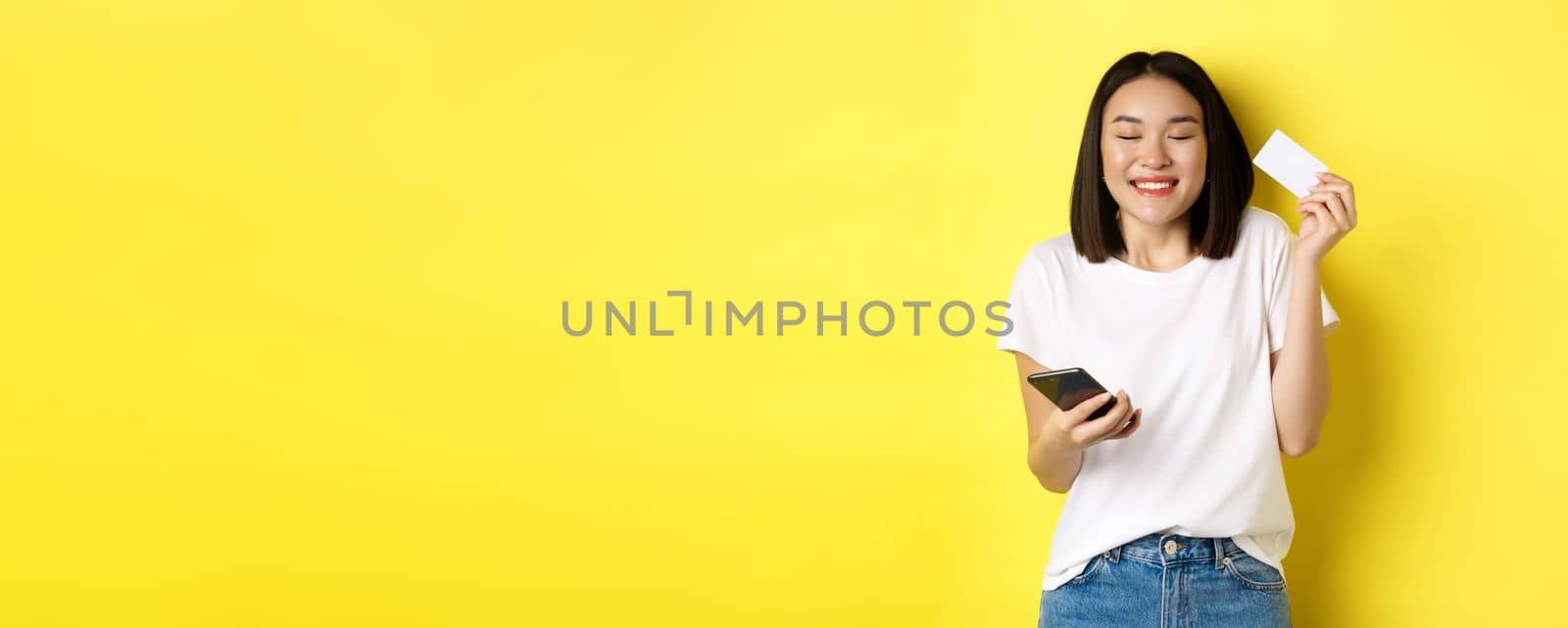 E-commerce and online shopping concept. Happy asian woman looking excited, buying something in internet, holding smartphone and showing plastic credit card, yellow background by Benzoix