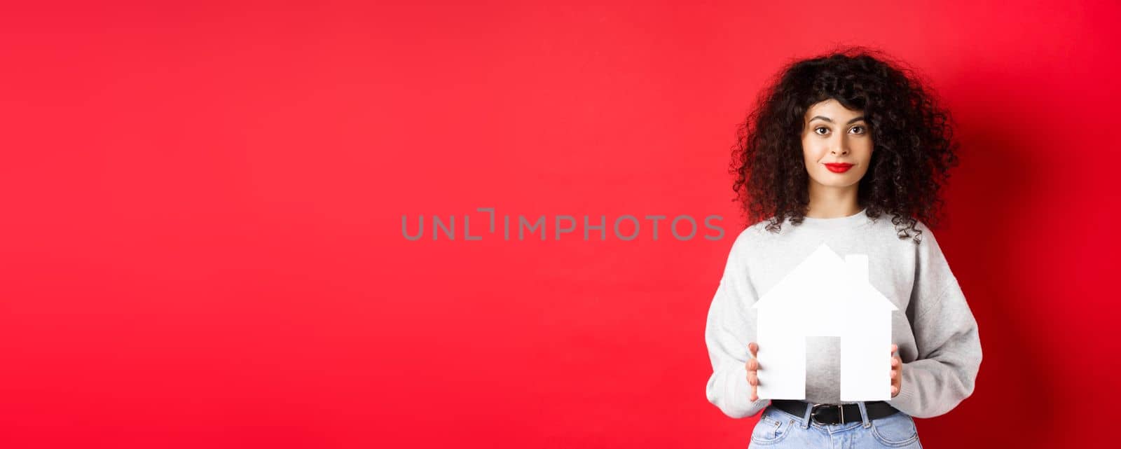 Real estate. Young caucasian woman in casual clothes showing paper house cutout, buying property or renting apartment, standing on red background by Benzoix