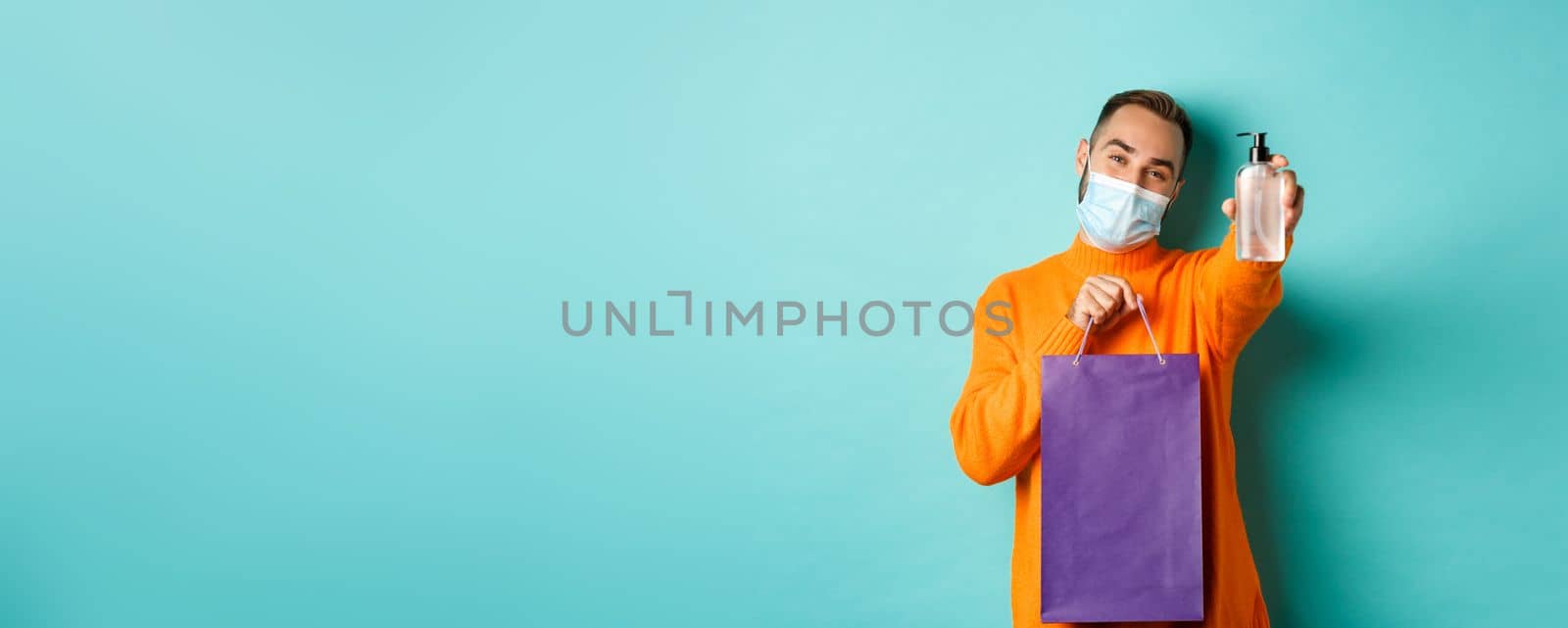 Coronavirus, pandemic and lifestyle concept. Man in face mask showing shopping bag and hand sanitizer, standing over turquoise background.