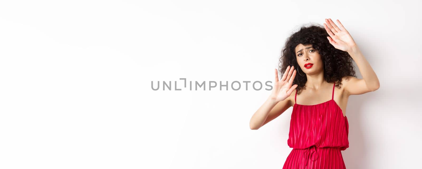 Young woman with curly hair and red dress, asking to stop, block someone, raising hands defensive, protecting herself, standing against white background.