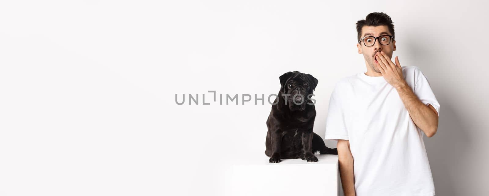 Image of dog owner and cute black pug looking at camera surprised and amazed, standing over white background by Benzoix