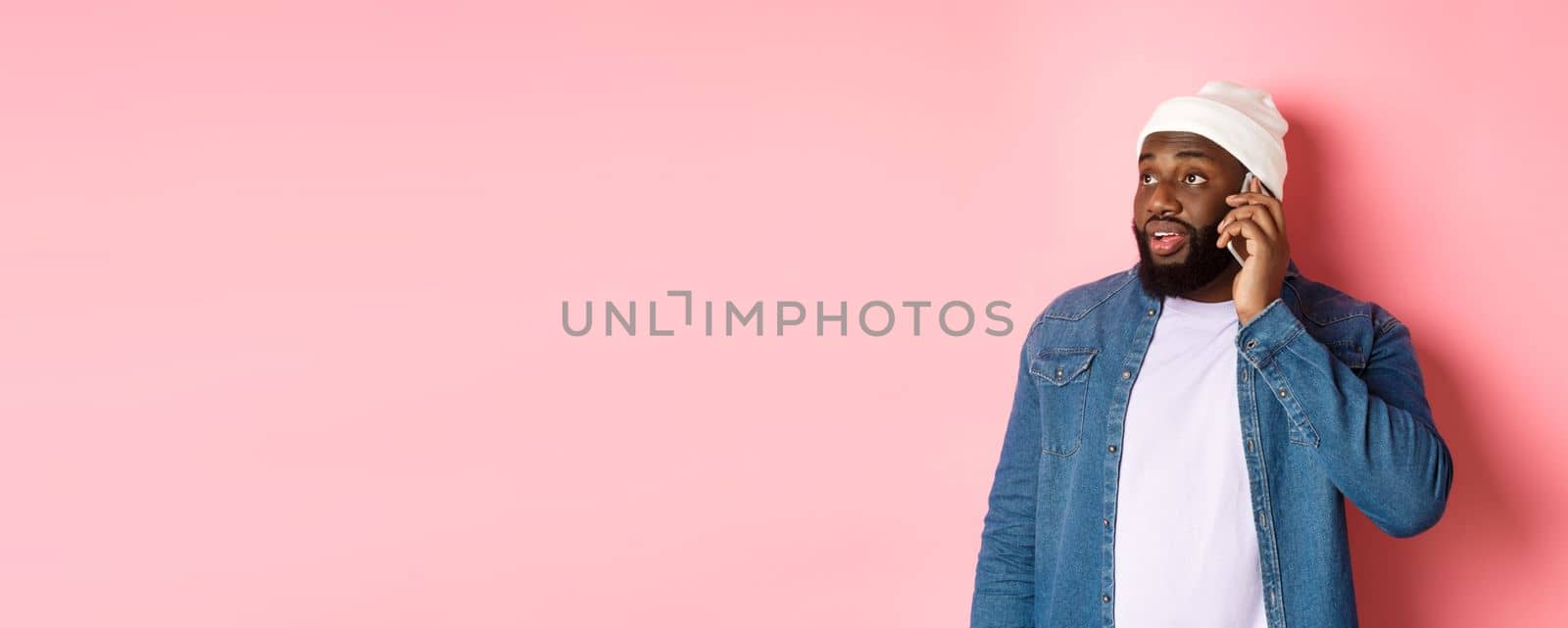 Hipster Black man talking on phone, looking left and having mobile conversation, standing over pink background.