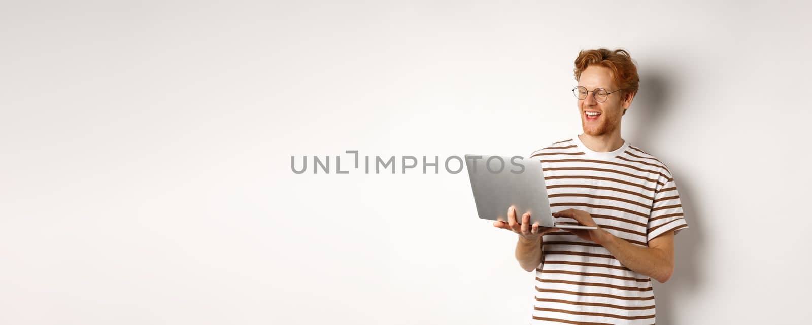 Handsome redhead man working freelance on laptop, smiling and looking pleased at computer screen, white background.